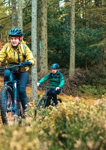 Two people on mountain bikes in woods, female in foreground, male in background.