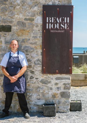 chef stood outside restaurant with beach in background.