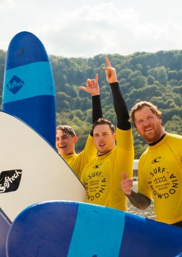 Team of four adult men raising their hands after surfing in the lagoon with yellow and black wetsuits on and blue surf boards.