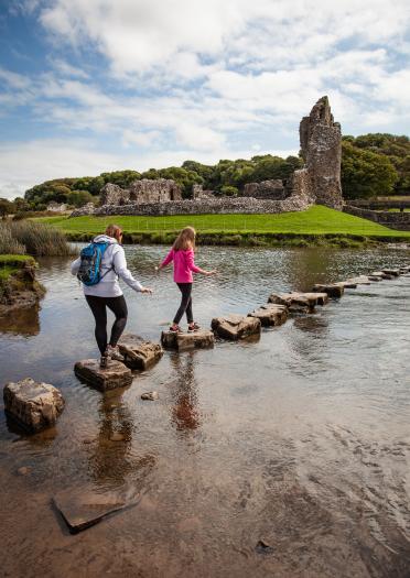 two young females walking on stepping stones with castle ruins in the background.