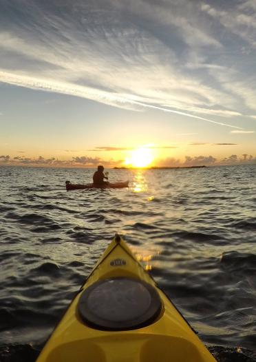 The front of a  yellow Kayak facing a person in another kayak with the sun setting in the background.