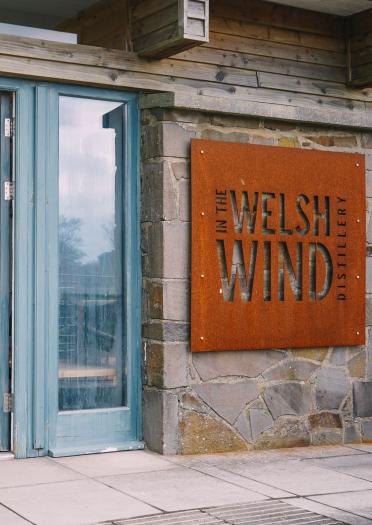 exterior of building with sign In the Welsh Wind and open doors showing interior with drinks.