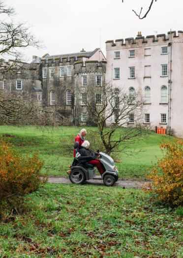 A lady in a motorised buggy on a path in front of a castle.