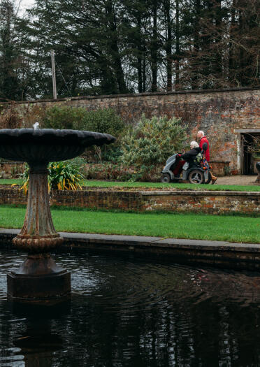 A woman in a motorised scooter and a man walking in an ornamental garden.
