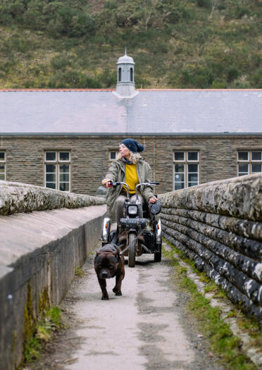 A lady in a mobility scooter and her dog crossing over a stone bridge.