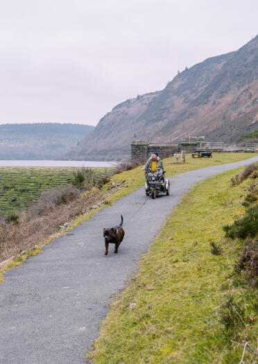 Eine Frau mit Elektromobil zusammen mit Hund auf einem geteerten Weg an einem Stausee.