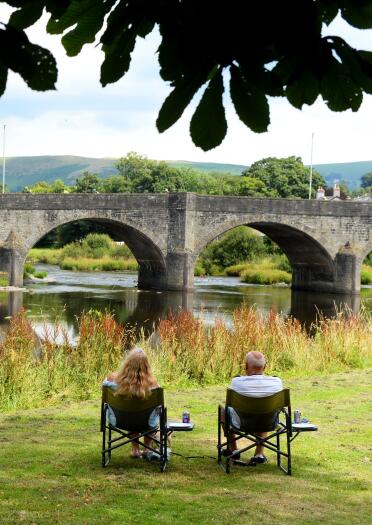 Two people in picnic chairs in front of a stone bridge spanning a wide river.