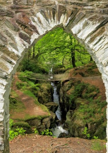 A woodland waterfall and chain bridge seen through a gothic arch.