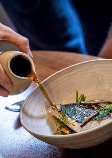 A person pouring a broth onto a fish dish in a bowl.