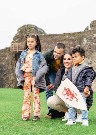 man, woman and two children on grass, boy with shield and castle wall in background.