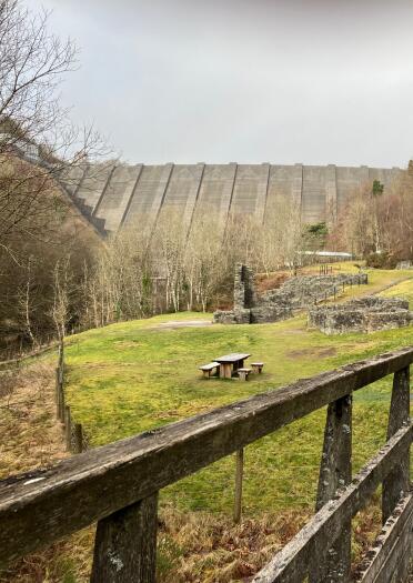 Ruins of industrial buildings on grassy land, under a high dam. 