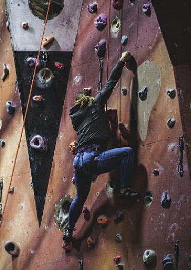 Climbing harnessed female high on indoor red climbing wall in blue sports trousers and hoodie.