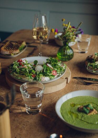 Meals on a wooden table. There is a bowl of-green soup and a plate of salad. There is also white wine and water on the table.