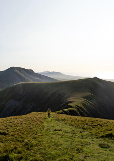 A person walking along a grassy mountain top path towards more mountains.