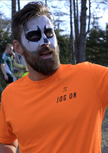 A bearded man with ghost make up on, wearing a bright orange top, in a forest. 