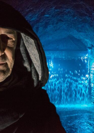 man with hood in foreground with cave and water light up in blue in the background .