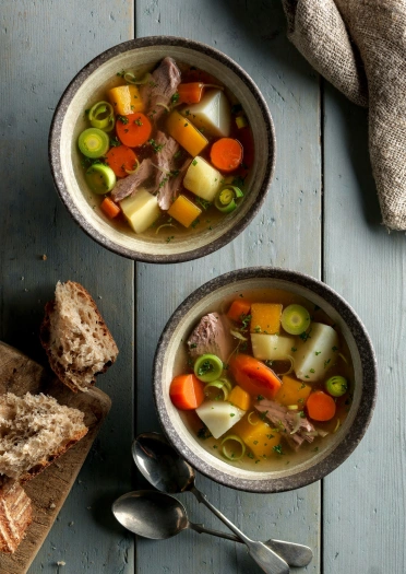 two bowls of cawl with bread and spoons on wooden table.