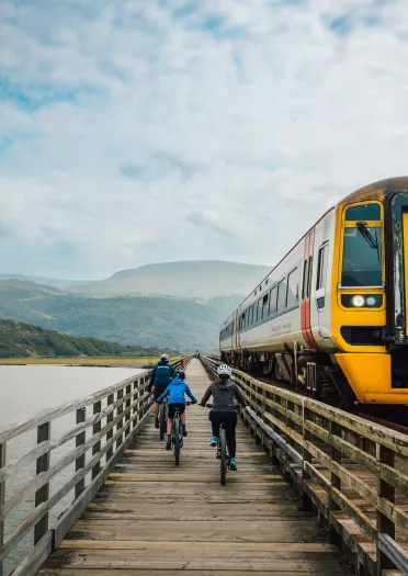 Three cyclists on a wooden railway bridge path, with a train passing by.