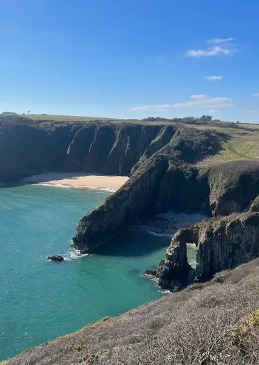 Coastal cliff landscape with a sandy beach and turquoise water under a clear blue sky. Sparse vegetation covers the cliffs and hills in the background.