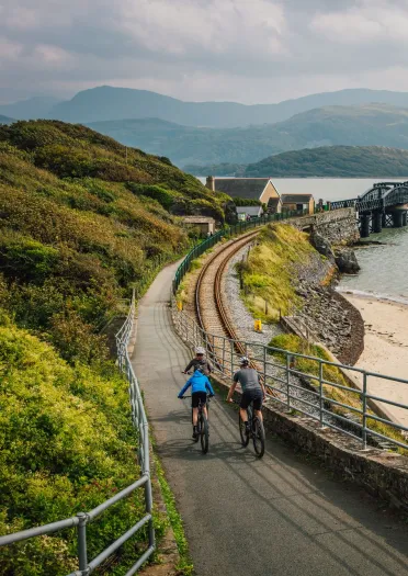 Drei Radfahrer fahren über eine breite Holzbrücke, die über einen Fluss führt, im Hintergrund sind grüne Hügel und bewaldete Hänge zu sehen.