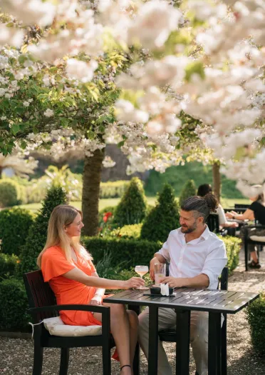 Two people seated at an outdoor table in a lush garden, engaged in conversation amid blooming trees and greenery