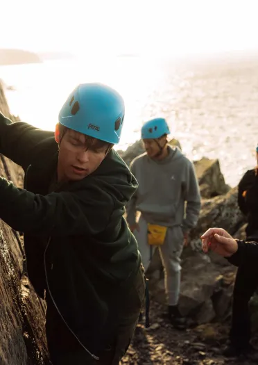 climber with helmet reaching up to rockface guided by instructor with people wearing helmets watching.