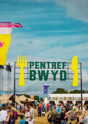 Sign in Welsh reading 'pentre bwyd' with large fork and knife, crowds of people and food tents.