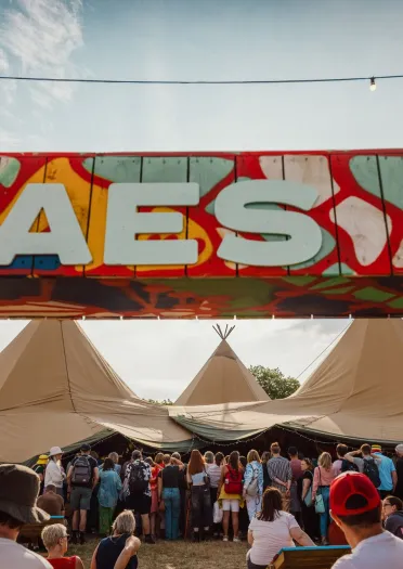 Sign reading 'Maes b' with festival tents and people.
