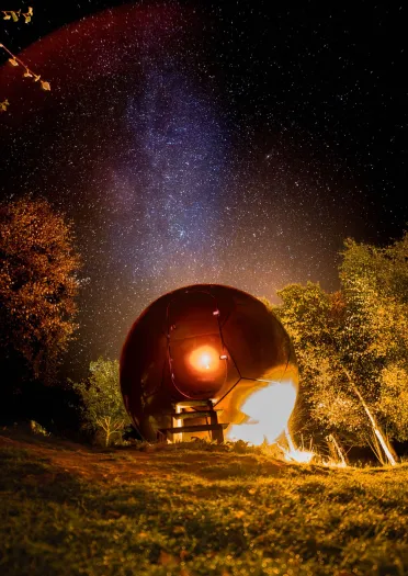 Illuminated dome-shaped structure at night, set in a grassy area with surrounding trees and a starry sky in the background.