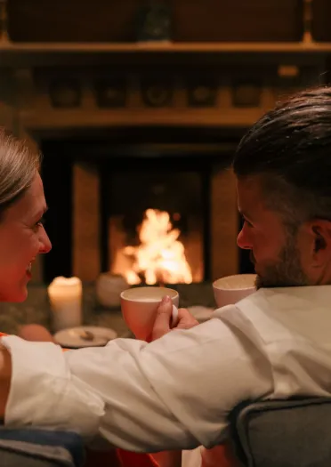 Couple seated on a couch by a lit fireplace, holding mugs and surrounded by candlelight in a cozy indoor setting.