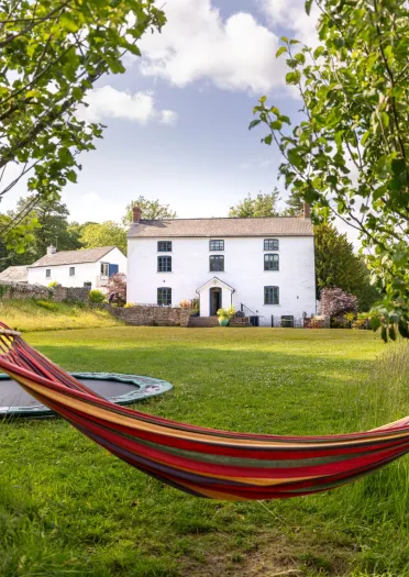 Colourful hammock strung between two trees in a grassy garden, with a white farmhouse and trampoline in the background under a blue, partly cloudy sky