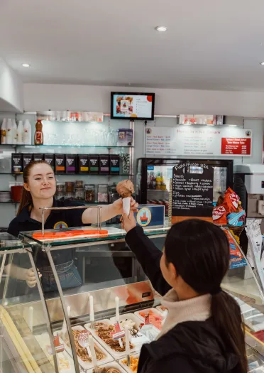 A customer reaches across an ice cream counter to receive a cone from a staff member inside a brightly lit shop displaying various flavours and toppings.