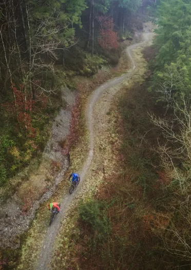 Aerial view of two cyclists riding along a winding dirt trail through a steep, forested valley with tall evergreens on one side and rocky terrain on the other.