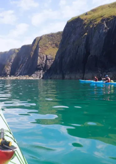 A kayaker’s viewpoint across calm turquoise water towards steep rocky cliffs, with two people paddling a blue kayak near the shoreline.
