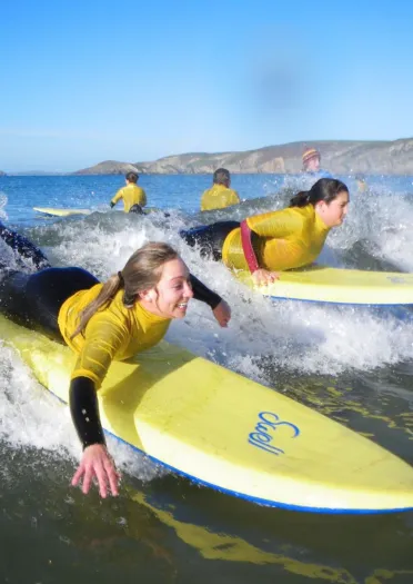A group of people lying on yellow surfboards catching small waves close to shore, wearing matching yellow tops with hills visible in the background under a clear blue sky.