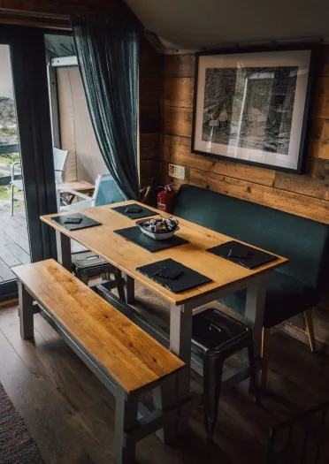 Wood‑panelled glamping cabin dining area with a table, bench seating, and doors opening onto a deck with views of the surrounding landscape.