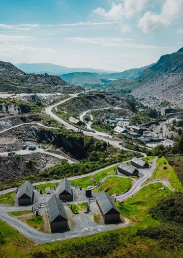 Aerial view of a small cluster of glamping cabins set in a mountainous quarry landscape, with winding roads and hills in the background.