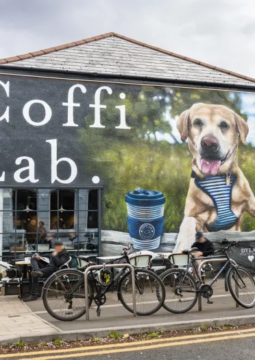 Exterior view of Coffi Lab café with outdoor seating and bicycles parked outside, featuring a large wall mural of a dog beside a takeaway coffee cup.