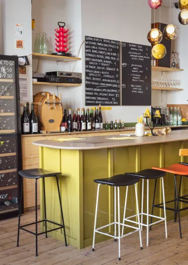 A small bar with a yellow counter, wooden stools and shelves of bottles along the wall.