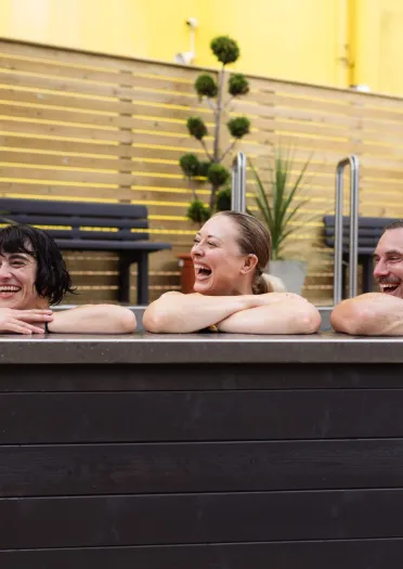 Three people leaning on the edge of an outdoor heated pool, with a “30+ Temperature” sign on the pool wall and wooden fencing and plants behind.