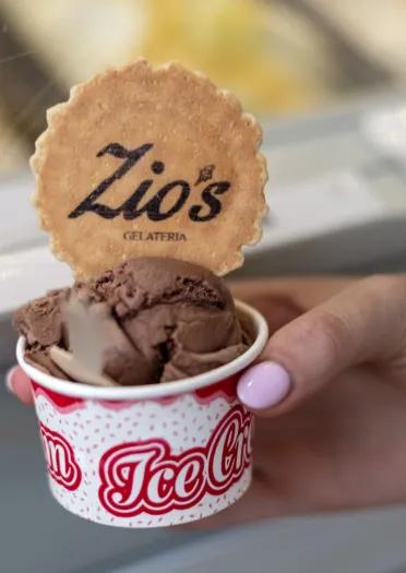 Hand holding a cup of chocolate gelato topped with a branded biscuit at Zio’s Gelateria