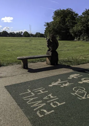A bench with a wooden sculpture at one end, in a city park next to a shared pathway..
