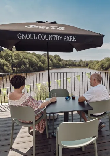 Visitors seated on an outdoor terrace overlooking the lake at Gnoll Country Park, with woodland reflected in the water below.
