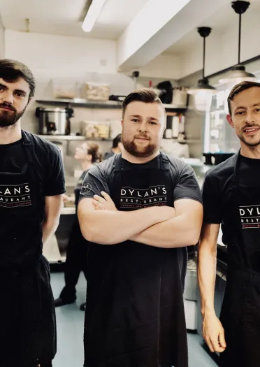 Three members of staff wearing black Dylans Restaurant aprons standing in a commercial kitchen, with stainless steel equipment, shelves and hanging lights in the background.