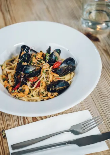 Seafood pasta with mussels, prawns and red pepper served in a white bowl on a wooden table, with cutlery and a glass of water alongside.