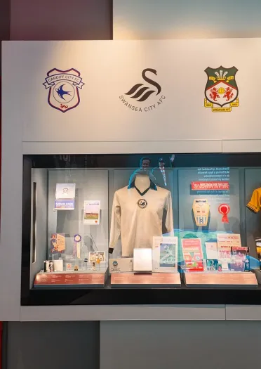 Display case at the Football Museum of Wales featuring memorabilia, historic football shirts, programmes and artefacts from Cardiff City, Swansea City, Wrexham AFC, Newport County and the Wales national team.