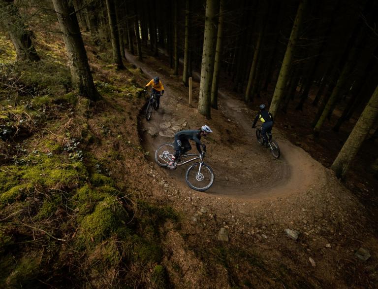Three mountain bikers going around a curved dirt track in a forest.