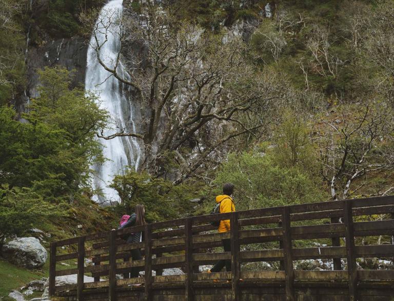 People walking over a footbridge over a stream looking up at a waterfall.