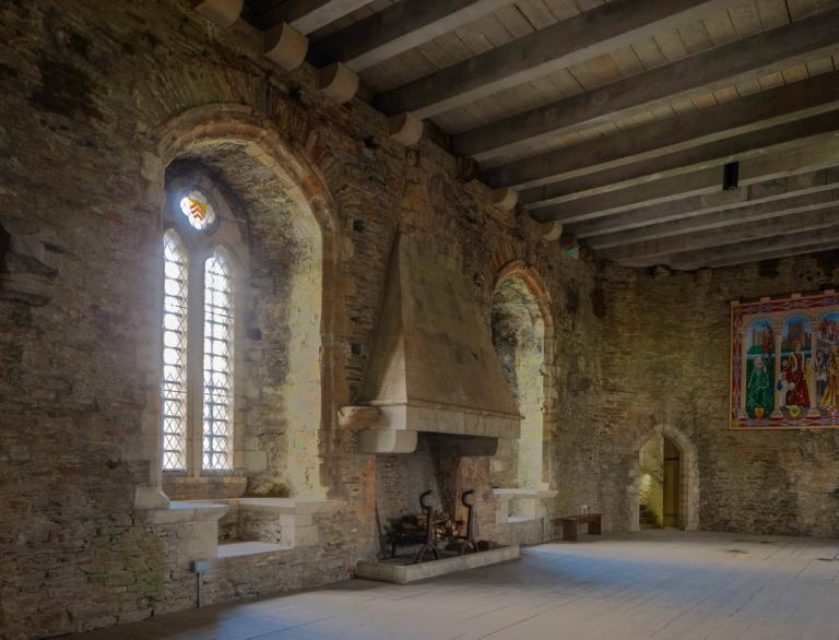 A room in a castle with large arched windows, fireplace and tapestry.