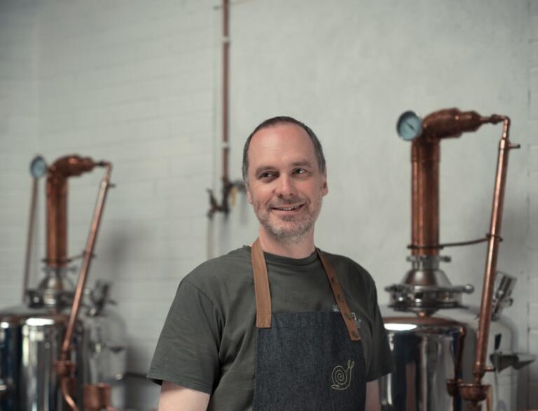 Mann in Schürze und Hemd vor einer handwerklich betriebenen Gin-Destillerie.